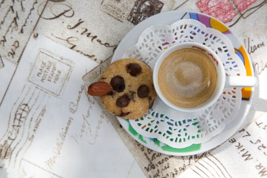 A cup of espresso and a shortcake with chocolate chips and almond nut on the letters print table cloth. Top view.