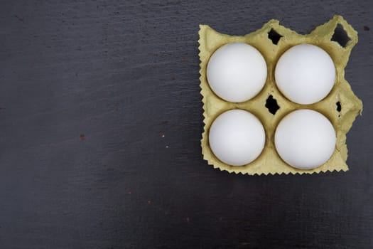 Four white chicken eggs in the carton box on the black wooden surface. Free space for a text.