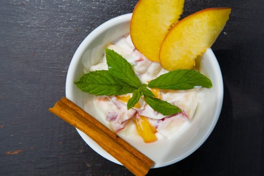 Homemade peach yogurt in a white dish on the black wooden surface. Decorated with two slices of fresh peach and fresh peppermint leaves and cinnamon stick. Top view. 