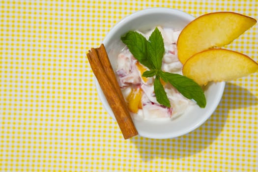 Homemade peach yogurt in a white dish on a yellow table cloth. Decorated with two slices of fresh peach and fresh peppermint leaves and cinnamon stick. Top view. 