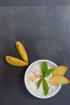 Homemade peach yogurt in a white dish on the black wooden surface. Decorated with two slices of fresh peach and fresh peppermint leaves. Top view. Free space for a text