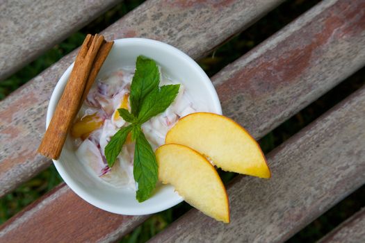 Homemade peach yogurt in a white dish on the old wooden surface. Decorated with two slices of fresh peach and fresh peppermint leaves and cinnamon stick. Top view. 
