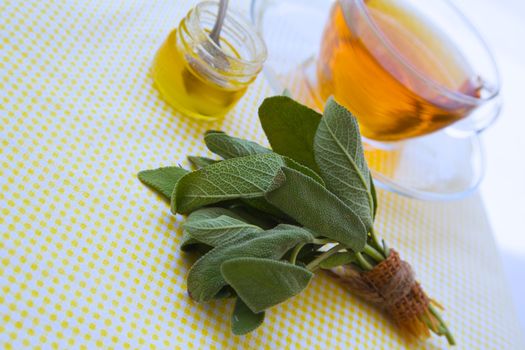 Fresh leaves of garden sage on a yellow table cloth. Tea and honey glass in the background