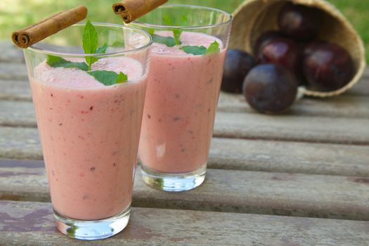 Two glasses of plum smoothie with cinnamon. Decorated with fresh peppermint leaves and cinnamon sticks. Fresh plums in a woven basket in the background