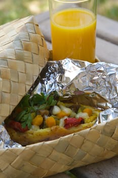 Open-air lunch for a vegetarian- potato baked with vegetables and cheese in an aluminium foil and a glass of fresh orange juice. Dish is hidden in the woven birch basket