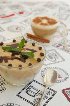 Pudding with cappuccino flavour in a glass dish. Decorated with coffee beans ,chocolate topping and fresh peppermint leaves. A cup of cappuccino in the background