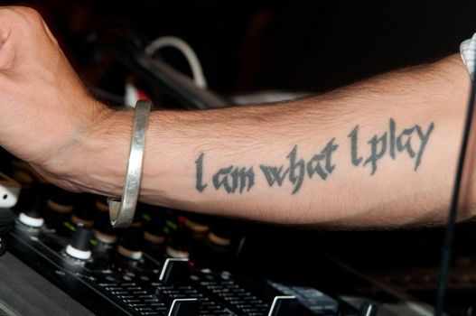 A man shows his bracelet and his tattoo saying I am what I play (likely a dj)