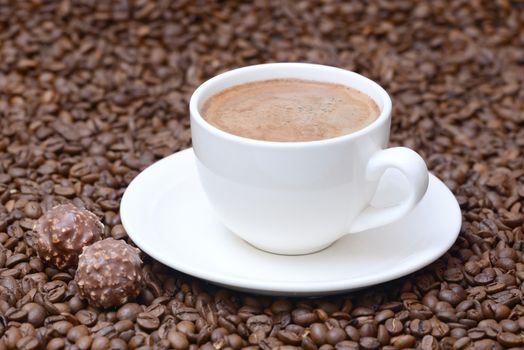 Photo of a cup of coffee on a saucer with candies. White color cup on a brown coffee beans background. Food photography.