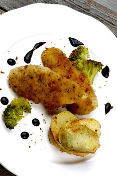 Delicious Fried Chicken Strips in Herb Breading Served with Steamed Broccoli, Roasted Potato Slices and Balsamic Sauce on White Plate Cross Section on Rustic Wooden background