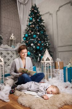 Young woman reading book to toddler son beside Christmas tree, boy falling asleep