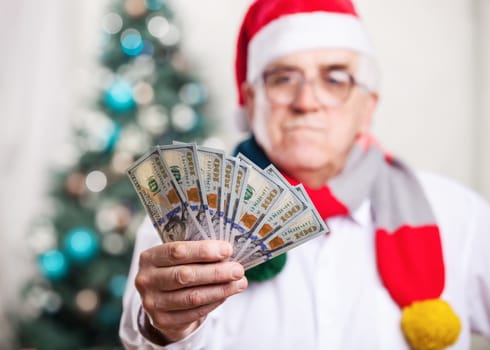 Senior man in Santa's hat holding money on Christmas background, hand in focus