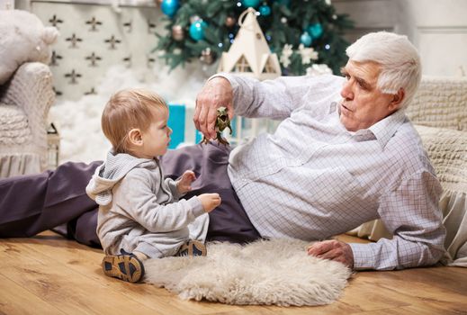 Toddler boy and his grandpa playing with toy dinosaur at Christmas tree