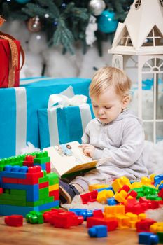 Cute toddler boy sitting at Christmas tree and reading book. Building blocks scattered around.