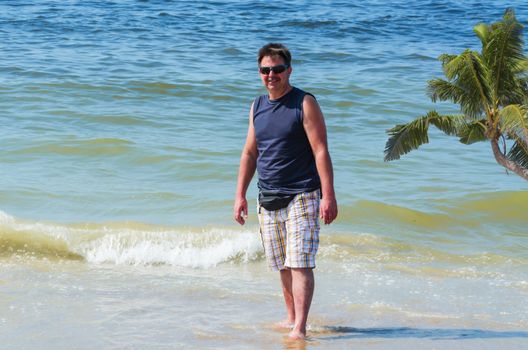 Man in shorts, T-shirt and sunglasses on the beach with your feet in the water.