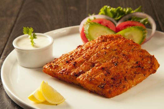 Grilled fish tikka served on a plate with salad and tarter sauce dip and salad with lemon, on a dark wood background