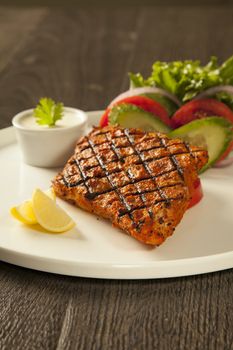 Grilled fish tikka served on a plate with salad and tarter sauce dip and salad with lemon, on a dark wood background