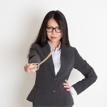 Asian female teacher holding a stick pointing at camera, standing on plain background.