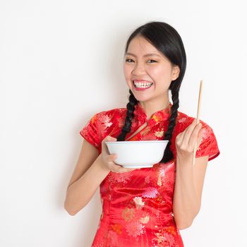 Portrait of Asian Chinese female eating, using chopsticks holding rice bowl, in traditional dress red cheongsam standing on plain background.