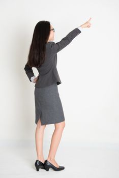 Full body backside Asian business woman pointing at copy space, standing on plain background.