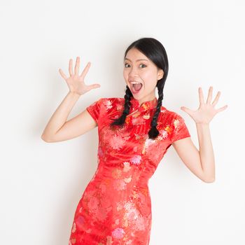 Portrait of Asian Chinese girl with surprised facial expression, in traditional red qipao standing on plain background.