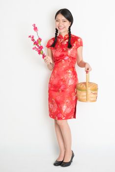 Portrait of full length Asian Chinese girl hands holding gift basket and plum blossom, in traditional red cheongsam standing on plain background.