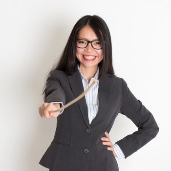 Asian female teacher smiling and holding a stick pointing at camera, standing on plain background.