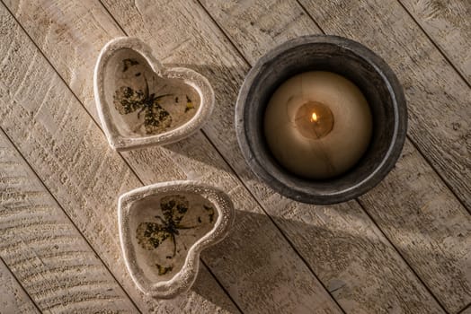 Two heart decorations on a white whashed wooden table next to a lit candle in a  stoneware pot all in low directional light of the late afternoon.