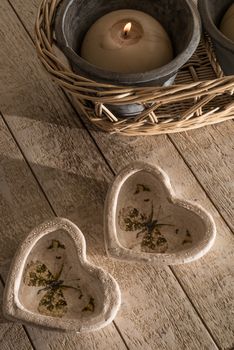 Two decorative hears  made from stone ware in the shape of little cups  or holders on a white whashed wooden table next to a burning ball candle in a pot,  and which in it's turn is inside a wicker basket.