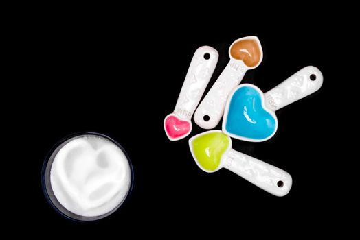 Heart shaped measuring spoons next to a small bowl of sugar with a hard shape inside the sugar, all on an isolated black background.