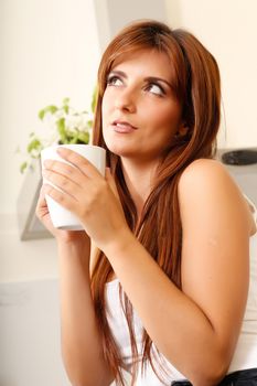 A young adult woman sitting in the kitchen with a cup of coffee. 