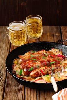 Sausages fried in cast iron skillet with two beer mugs on wooden background