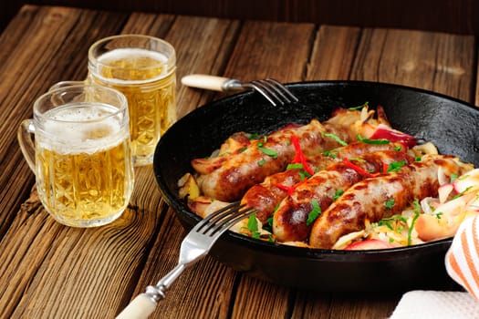 Sausages fried in cast iron skillet with two beer mugs on wooden background