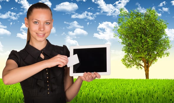 Beautiful businesswoman holding blank tablet PC and blank business card in front of PC screen. Green meadow, sky and tree as backdrop