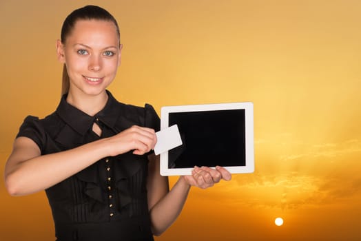 Beautiful businesswoman holding blank tablet PC and blank business card in front of PC screen. Bright sky at sunset as backdrop