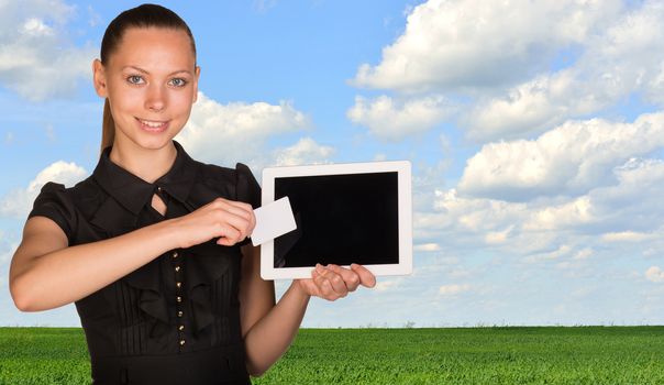 Beautiful businesswoman holding blank tablet PC and blank business card in front of PC screen. Green meadow, sky and clouds as backdrop