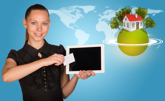 Beautiful businesswoman holding blank tablet PC and blank business card in front of PC screen. Green planet surrounded by horisontal rings, with house and trees on top, and world map as backdrop