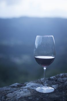 Crystal glass of red wine on stone wall in silhouette with sky and couds behind defocused. Monochrome blue grey tone artistic color digital photograph in Lucca Tuscany Italy in summer. 