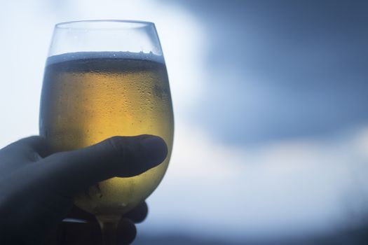 Hand of man holding glass of lager beer set against blue grey sky. Artistic color digital photo with shallow depth of focus.