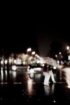 Defocused people crossing road carrying umbrellas in rain at night. Artistic color digital rectangular vertical photo with black background. 