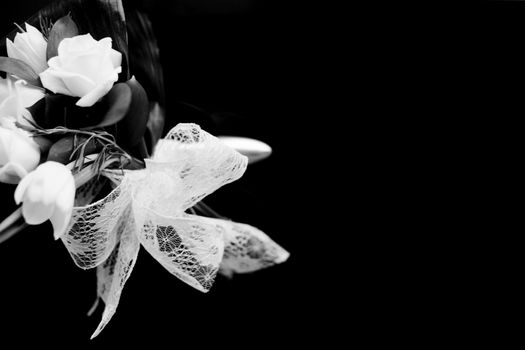 Black and white artistic digital rectangular horizontal photo of the bride's bridal bouquet of flowers with lace bow including white white roses before marriage ceremony in Spain. Shallow depth of with plain black background out of focus. 