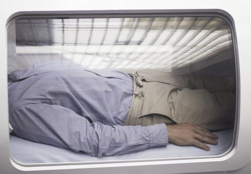 Male patient aged 45-55 wearing blue shirt and beige chino trousers lying down in hyperbaric oxygen chamber receiving Hyperbaric Oxygen Therapy (HBOT) specialised medical treatment for injuries with reflection on glass. 
