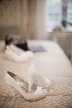 Color artistic digital rectangular vertical photo of the bride's white high heel shoes on bed in luxury hotel suite bedroom. Shallow depth of focus with background defocused.