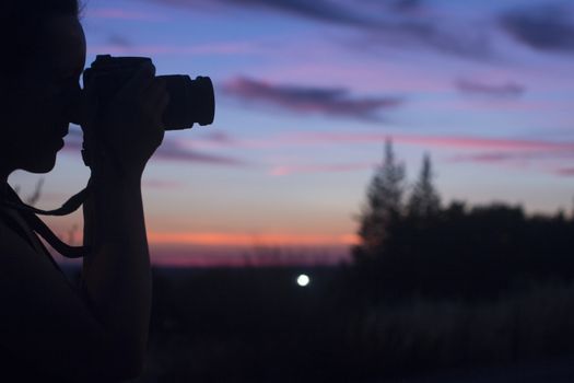 Female photographer taking photo in silhouette against blue purple sky with medium format analogue film camera and strap with long lens and filter holder on hot summer evening. 