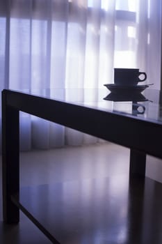 Artistic photo of coffee or tea cup with saucer and tea spoon on a glass table with reflection in room with window and net curtains behind in soft evening purple blue color light out of focus in background. 
