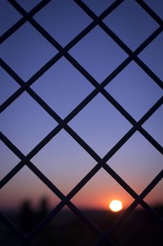 Color digital photo of setting evening sun and red and blue sky shining through a metal wire mesh fence in silhouette at dusk in dark tones with bokeh blur shallow depth of focus defocused landscape background. 