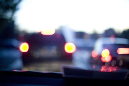 Road at dusk sunset seen from inside a car driving in traffic with other cars and rear lights in bokeh out of focus with shallow depth of focus on dashboard.