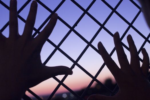 Color digital photo of setting evening sun and red and blue sky shining through a metal wire mesh fence with two human hands of a man against the fence with outstretched fingers in silhouette at dusk in dark tones with bokeh blur shallow depth of focus defocused landscape background. 