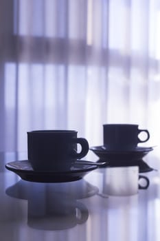 Artistic photo of coffee or tea cup with saucer and tea spoon on a glass table with reflection in room with window and net curtains behind in soft evening purple blue color light out of focus in background. 
