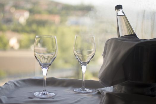 Still life photo of two glasses and bottle of water in metal icebucket with cloth covering on a table in room with window behind in soft evening pastel color light out of focus in background. 