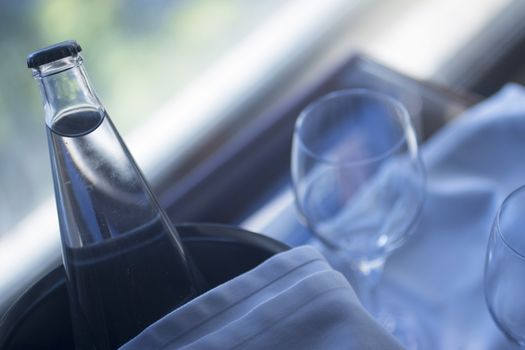 Artistic still life photo of two glasses and bottle of water in metal ice bucket with cloth covering on a glass table in room with window behind in soft evening purple blue color light with out of focus in background.